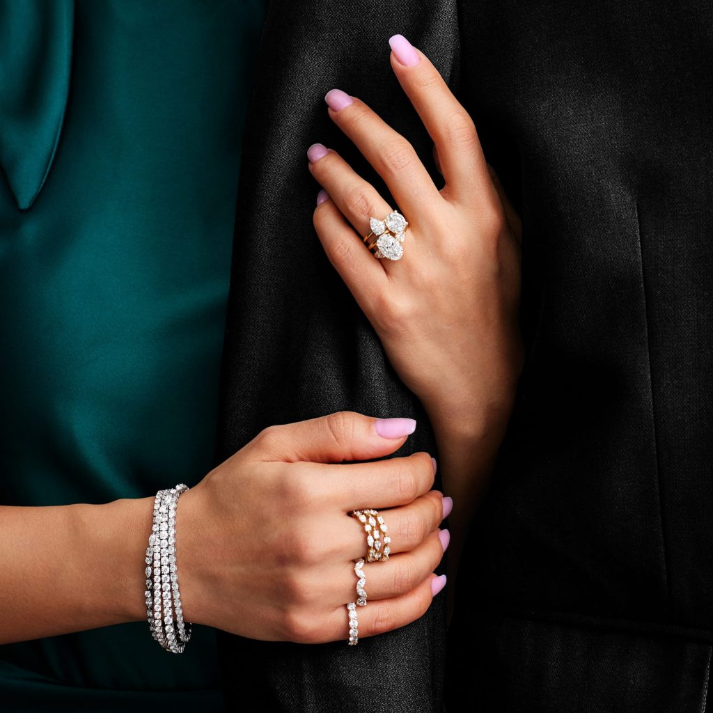 A close-up of a woman's hands adorned with sparkling diamond rings and a multi-row diamond bracelet, resting against a dark coat and emerald-green fabric.